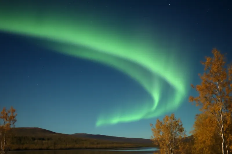 Aurore boréale verte au-dessus d'un lac lapon bordé de bouleaux dorés, sol encore libre de neige en septembre